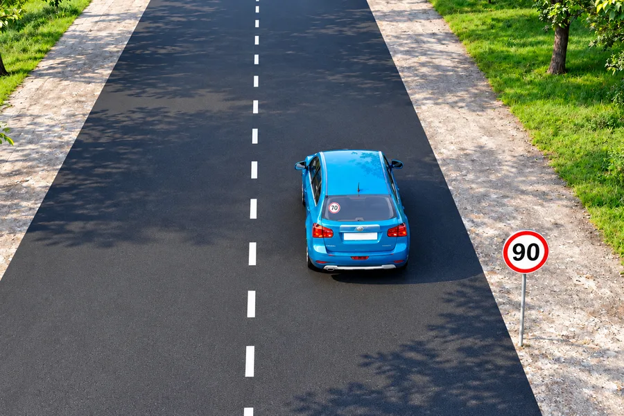 Orqa oynasida 70 taniqlik belgisi bor avtomobil, yo'l chetida 90 km/soat belgisi